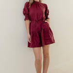 Woman wearing a burgundy mini dress with high neck, short puff sleeves, and a tiered skirt, styled with waist tie.