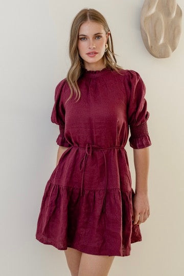 Woman wearing a burgundy mini dress with high neck, short puff sleeves, and a tiered skirt, styled with waist tie.