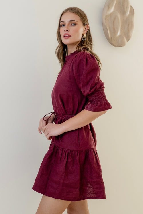 Woman wearing a burgundy mini dress with high neck, short puff sleeves, and a tiered skirt, styled with waist tie.