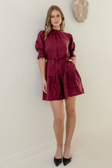 Woman wearing a burgundy mini dress with high neck, short puff sleeves, and a tiered skirt, styled with waist tie.