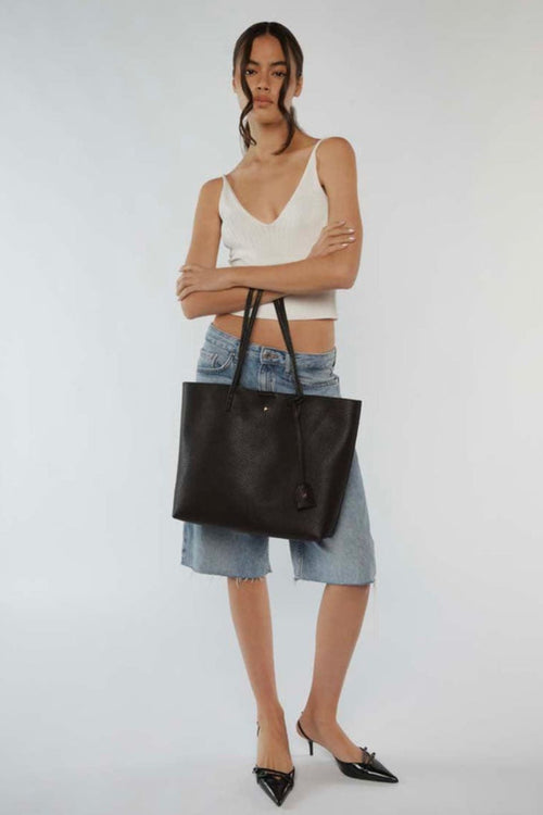 A sleek black pebble-textured vegan leather tote bag with long shoulder straps, minimal gold detailing, and a small matching tag charm hanging from one handle. The bag has a structured rectangular shape and a clean, minimalist design