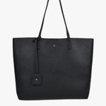 A sleek black pebble-textured vegan leather tote bag with long shoulder straps, minimal gold detailing, and a small matching tag charm hanging from one handle. The bag has a structured rectangular shape and a clean, minimalist design