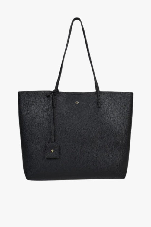 A sleek black pebble-textured vegan leather tote bag with long shoulder straps, minimal gold detailing, and a small matching tag charm hanging from one handle. The bag has a structured rectangular shape and a clean, minimalist design