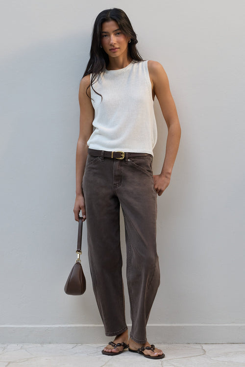 A woman stands against a light gray wall wearing a sleeveless white ribbed knit top, dark brown wide-leg pants, a brown belt, and brown sandals, while holding a small brown handbag.
