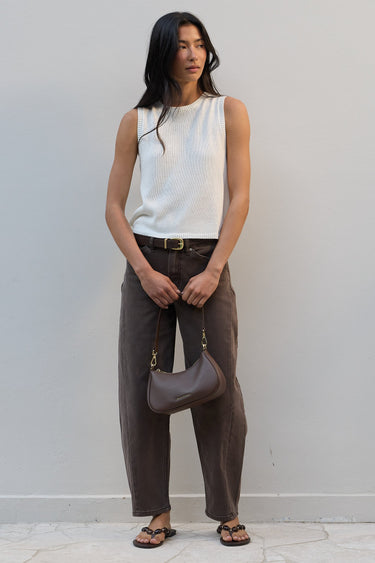 A woman stands against a light gray wall wearing a sleeveless white ribbed knit top, dark brown wide-leg pants, and brown sandals, while holding a taupe-brown crescent shoulder bag.