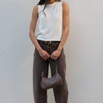 A woman stands against a light gray wall wearing a sleeveless white ribbed knit top, dark brown wide-leg pants, and brown sandals, while holding a taupe crescent-shaped shoulder bag.