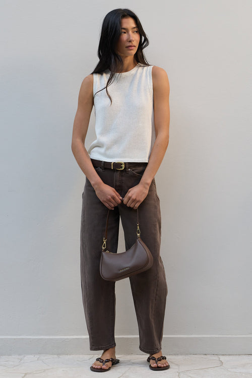 A woman stands against a light gray wall wearing a sleeveless white ribbed knit top, dark brown wide-leg pants, and brown sandals, while holding a taupe crescent-shaped shoulder bag.