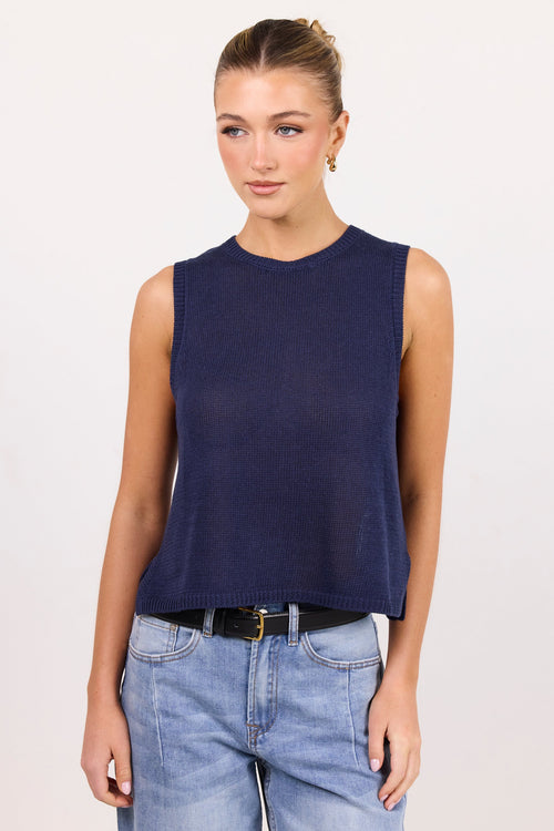 A woman modeling a sleeveless crewneck knit top in dark navy blue, paired with light blue jeans and a black belt, against a plain off-white studio background.