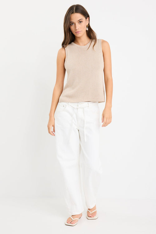 Woman wearing a sleeveless sand-colored knit tank top with a crew neckline and relaxed fit, paired with high-waisted white jeans.