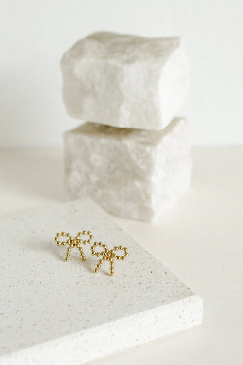 A pair of delicate gold-toned stud earrings shaped like bows, crafted from small polished metal beads arranged in a symmetrical design