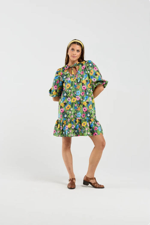 A woman modeling a short green floral dress with puff sleeves, a tie neckline, and a ruffled hem, paired with a yellow headband and brown sandals, against a plain light gray studio background.