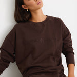 Relaxed-fit chocolate brown sweatshirt with tonal Re:Union embroidery, styled with cream shorts and gold hoop earrings.