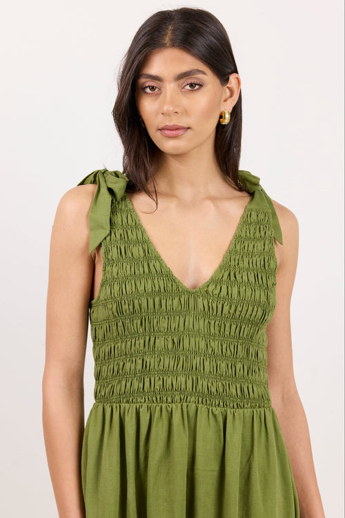 A woman modeling a sleeveless olive green dress with a smocked bodice, tie shoulder straps, and a gathered skirt against a plain light background.