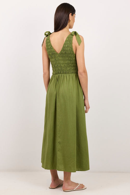A woman viewed from the back wearing an olive green sleeveless midi dress with shoulder ties and a smocked bodice, standing against a plain white studio background.