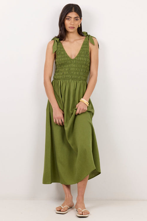 A woman modeling an olive green midi dress with a smocked bodice and tie shoulder straps, standing against a plain white studio background.
