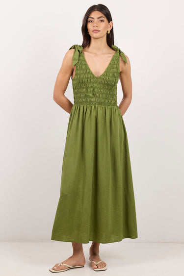 A woman modeling a sleeveless olive green midi dress with a smocked bodice and tie straps, standing against a plain white studio background.