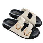 A pair of women's double-strap slide sandals by Stories Be Told, featuring off-white straps with gold-tone D-ring hardware and black soles, shown on a plain white background.