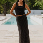 A model wears a long black skirt with an ivory geometric line print, featuring an elastic waist and a flowing silhouette, styled with a black sleeveless top and sandals.