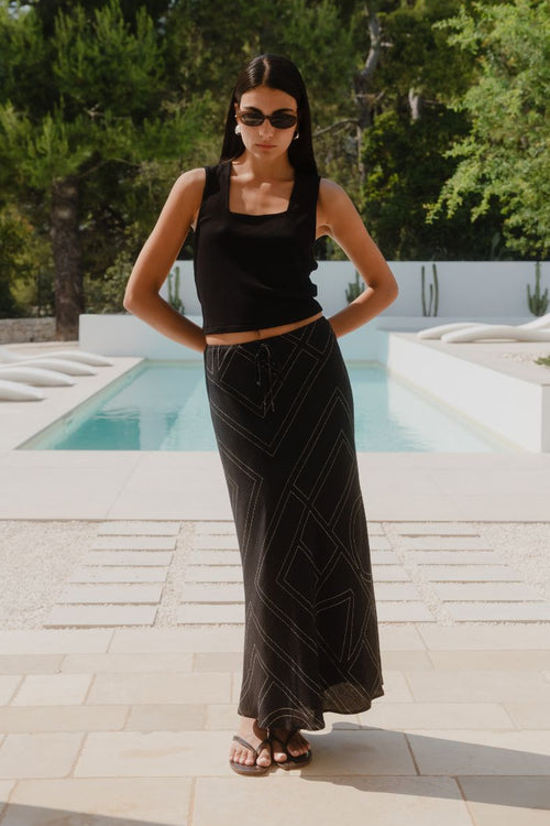A model wears a long black skirt with an ivory geometric line print, featuring an elastic waist and a flowing silhouette, styled with a black sleeveless top and sandals.