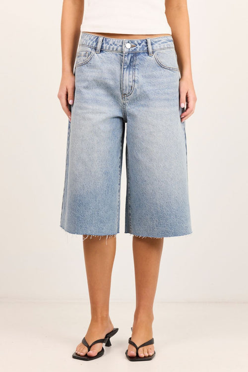 A model wearing light-wash blue denim cropped wide-leg jeans with a raw frayed hem, paired with black heeled sandals against a plain off-white studio background.