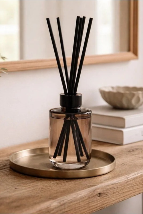 A reed diffuser with a smoky amber glass bottle and black reeds, displayed on a round brass tray on a wooden table in a softly lit interior setting.