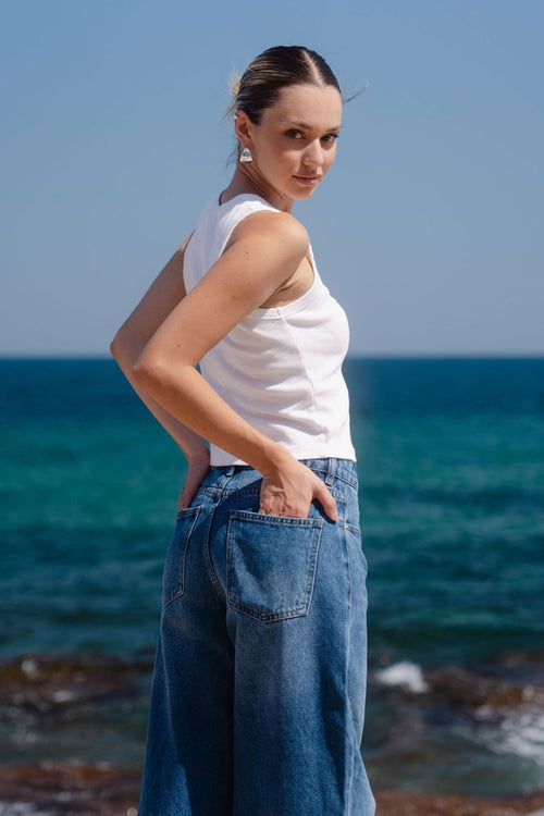 Woman standing on rocks by the ocean wearing a white top and blue jeans.