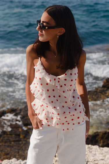 Woman wearing a cream cami top with cherry-red polka dots, ruffled front detailing, and thin adjustable straps