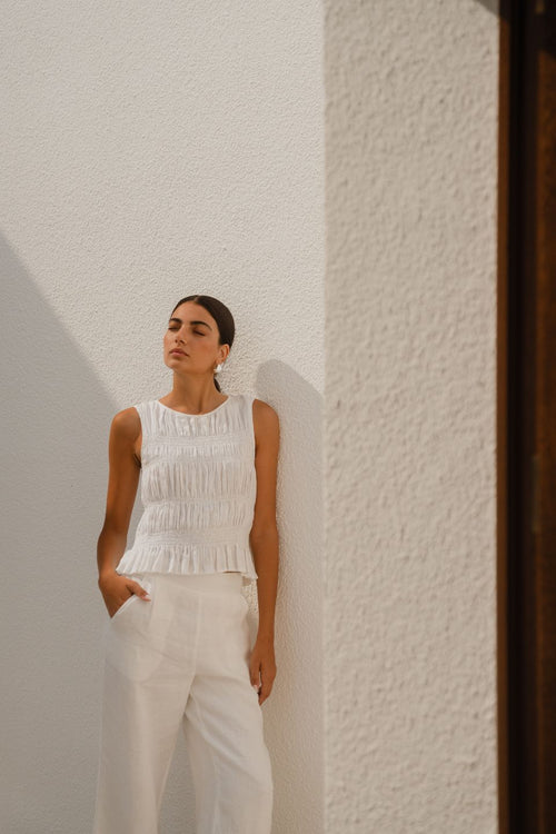 Woman wearing an ivory linen sleeveless top with a shirred bodice and ruffle hem, paired with dark blue jeans.