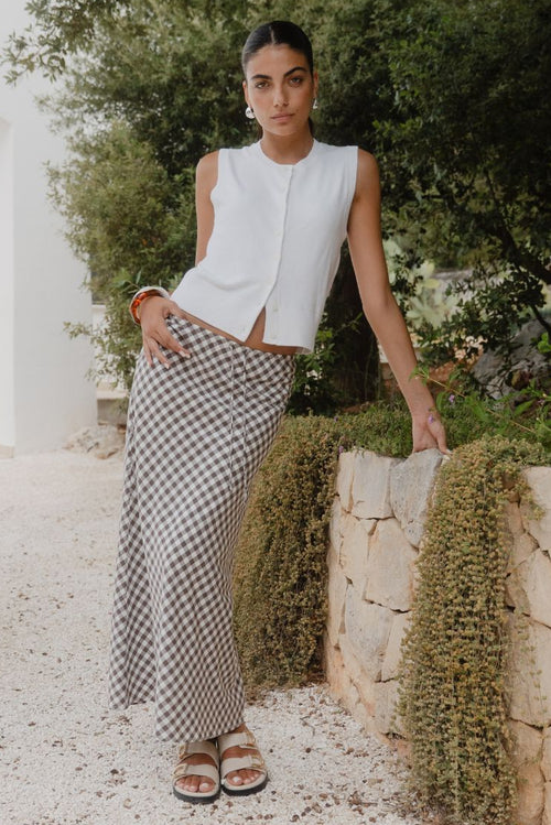 Woman in a white sleeveless top and checkered pants standing outdoors with greenery in the background