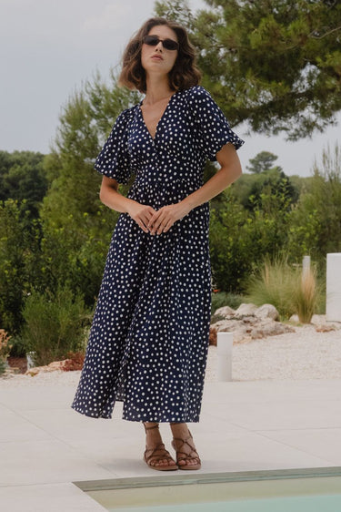 Woman in a navy blue dress with white polka dots standing by a pool with greenery in the background