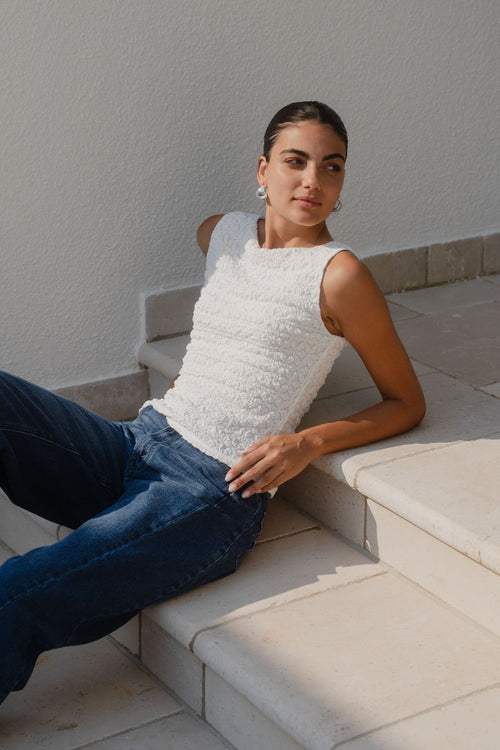 Woman wearing a white sleeveless textured top with a fitted silhouette and high neckline, paired with high-waisted denim jeans.