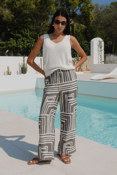 Woman wearing khaki and white print pants and a white top infront of a pool