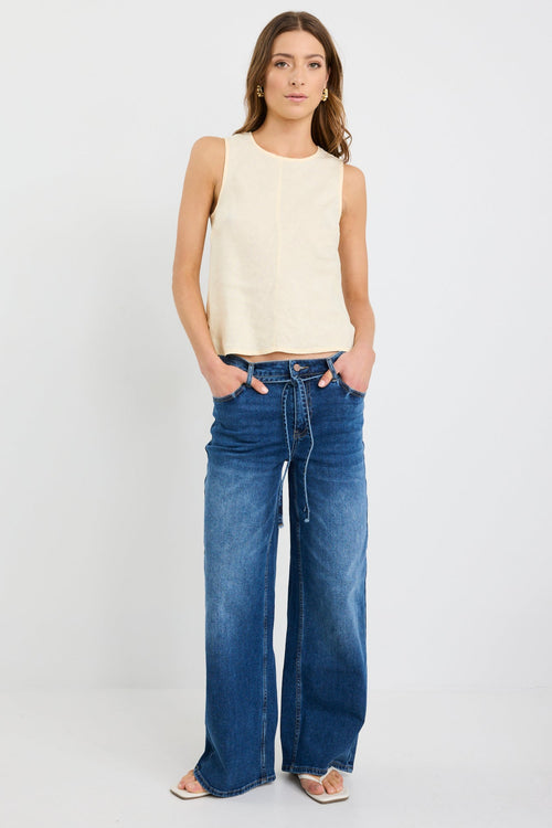 Woman wearing a sleeveless butter-yellow linen shell top with a round neckline and relaxed bias cut, styled with deep blue jeans.