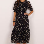 A woman modeling a black midi dress with a beige polka dot pattern, featuring short puff sleeves and a tiered skirt, set against a plain off-white studio background.
