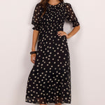 A woman modeling a black midi dress with a beige polka dot pattern, featuring short puff sleeves and a round neckline, set against a plain white studio background.