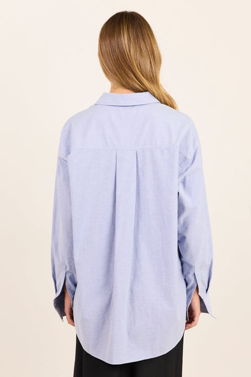 A woman viewed from the back wearing a light blue long-sleeve button-up shirt and black pants, standing against a plain cream-colored background.
