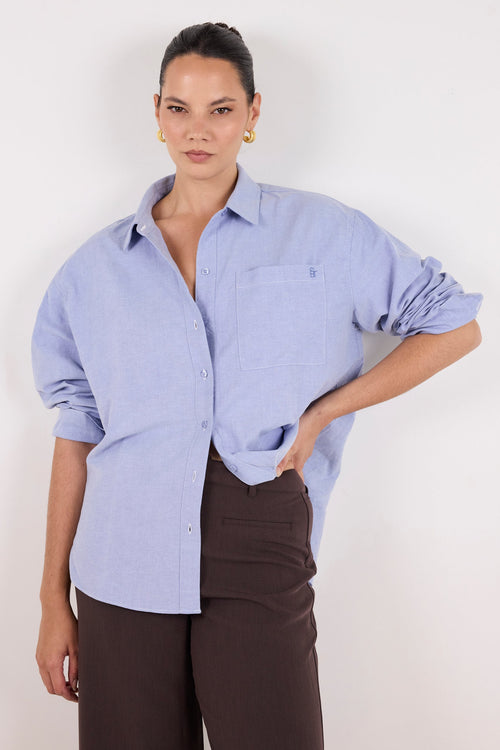 A woman modeling a light blue long-sleeve button-up shirt with rolled sleeves and dark brown trousers, standing against a plain off-white background.