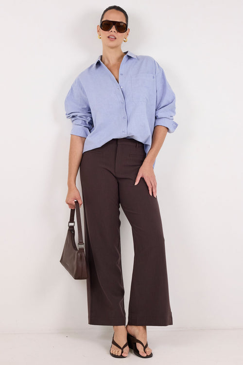 A model stands against a plain white studio background wearing a light blue button-down shirt, dark brown wide-leg trousers, black sandals, and holding a dark brown handbag.