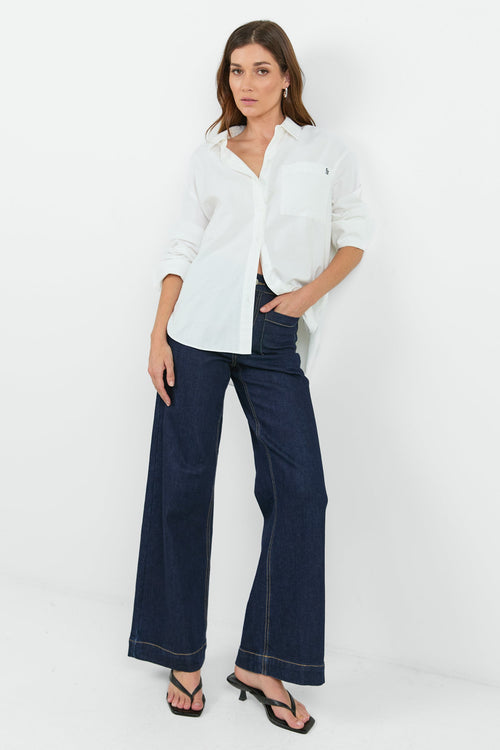 A woman wearing a classic white button-down shirt with a pocket on the left chest, paired with dark indigo wide-leg jeans. She is standing with one hand in her pocket, showcasing a relaxed and casual look.