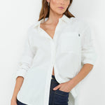 A woman wearing a classic white button-down shirt with a pocket on the left chest, paired with dark indigo wide-leg jeans. She is standing with one hand in her pocket, showcasing a relaxed and casual look.