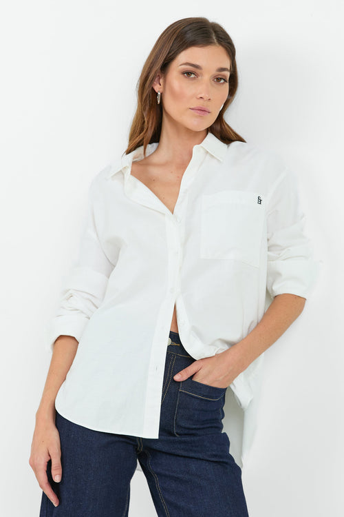 A woman wearing a classic white button-down shirt with a pocket on the left chest, paired with dark indigo wide-leg jeans. She is standing with one hand in her pocket, showcasing a relaxed and casual look.