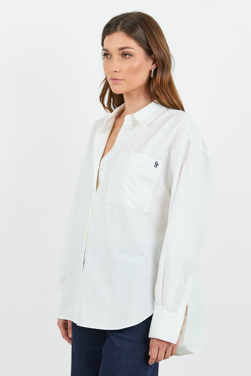 A woman wearing a classic white button-down shirt with a pocket on the left chest, paired with dark indigo wide-leg jeans. She is standing with one hand in her pocket, showcasing a relaxed and casual look.