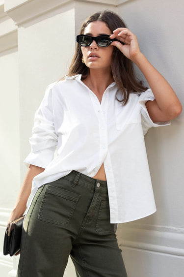 Woman in white shirt and olive green pants standing against a white wall.