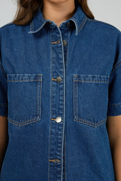 Woman wears a dark blue denim shirt with short sleeves and front patch pockets, styled with matching high-waisted denim shorts. The shirt is unbuttoned at the bottom for a relaxed, effortless look.