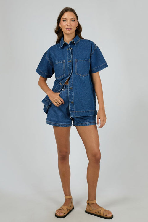 Woman wears a dark blue denim shirt with short sleeves and front patch pockets, styled with matching high-waisted denim shorts. The shirt is unbuttoned at the bottom for a relaxed, effortless look.