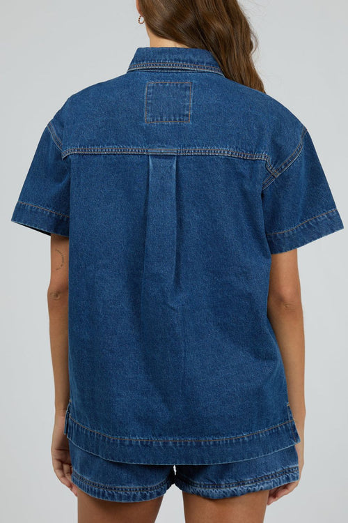 Woman wears a dark blue denim shirt with short sleeves and front patch pockets, styled with matching high-waisted denim shorts. The shirt is unbuttoned at the bottom for a relaxed, effortless look.