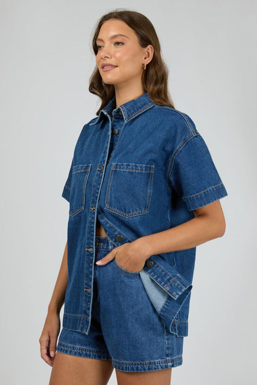 Woman wears a dark blue denim shirt with short sleeves and front patch pockets, styled with matching high-waisted denim shorts. The shirt is unbuttoned at the bottom for a relaxed, effortless look.