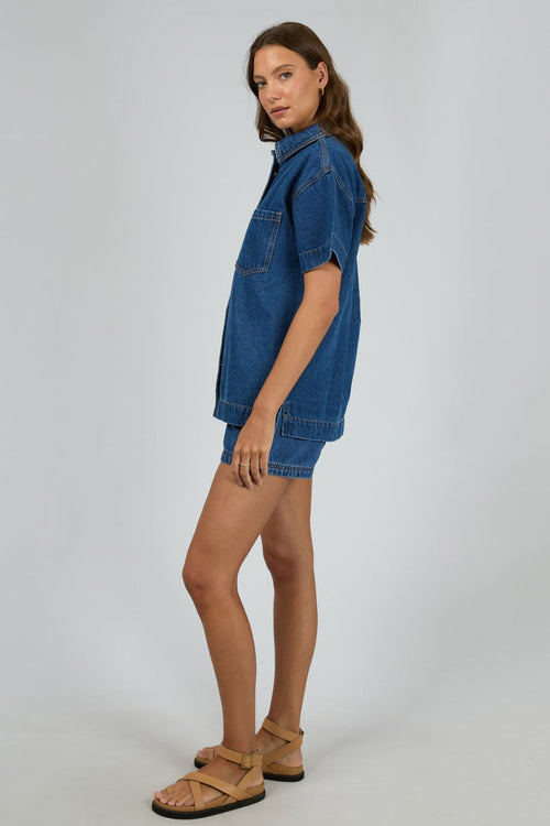 Woman wears a dark blue denim shirt with short sleeves and front patch pockets, styled with matching high-waisted denim shorts. The shirt is unbuttoned at the bottom for a relaxed, effortless look.