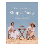 Book cover of 'Simple Fancy' by Two Raw Sisters with two women sitting at a table on a beach.