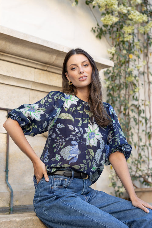 A woman wearing a navy blue floral puff-sleeve blouse and wide-leg blue jeans, sitting on stone steps outdoors with a beige wall and green ivy in the background.